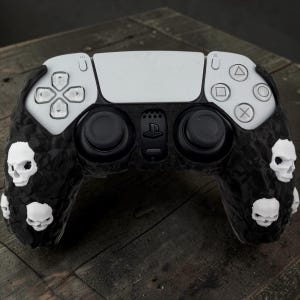 May include: A white and black video game controller featuring a textured black grip surface adorned with multiple small, white, three-dimensional skull accents. The controller includes standard directional buttons, two black analog sticks, and a central logo.