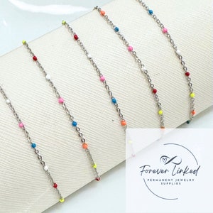 May include: Five silver chains with colorful beads. The chains are arranged in a row on a white surface. The beads are in various colors, including red, blue, green, yellow, and orange. The chains are all the same length and have a delicate, feminine look.