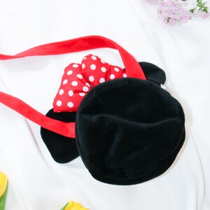 May include: Black plush handbag with a red strap and a red bow with white polka dots. The bag has two black ears. The bag is on a white background with yellow tulips and pink flowers.