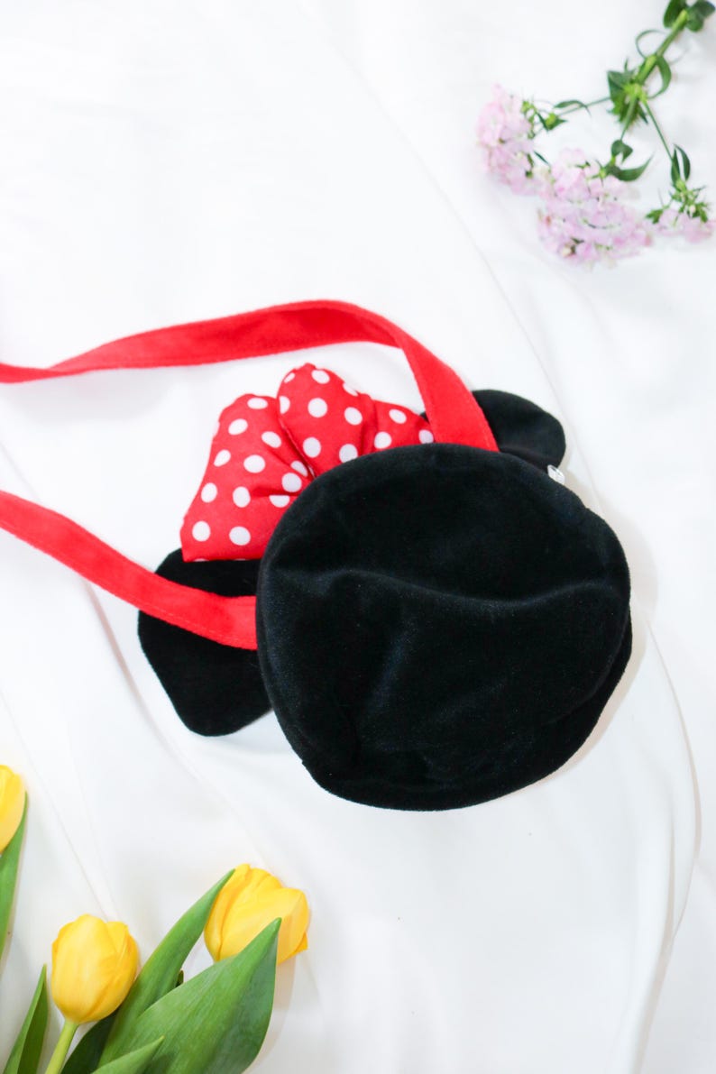 May include: Black plush handbag with a red strap and a red bow with white polka dots. The bag has two black ears. The bag is on a white background with yellow tulips and pink flowers.
