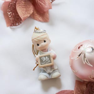 May include: A ceramic figurine of a child holding a chalkboard with the number 96. The figurine is wearing a crown and light blue overalls. The image also includes a pink Christmas ornament and decorative flowers.