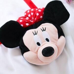 May include: A plush Minnie Mouse-shaped bag with a red strap. The bag features a pink face, black ears, and a red bow with white polka dots. The bag is designed to resemble the iconic Disney character.