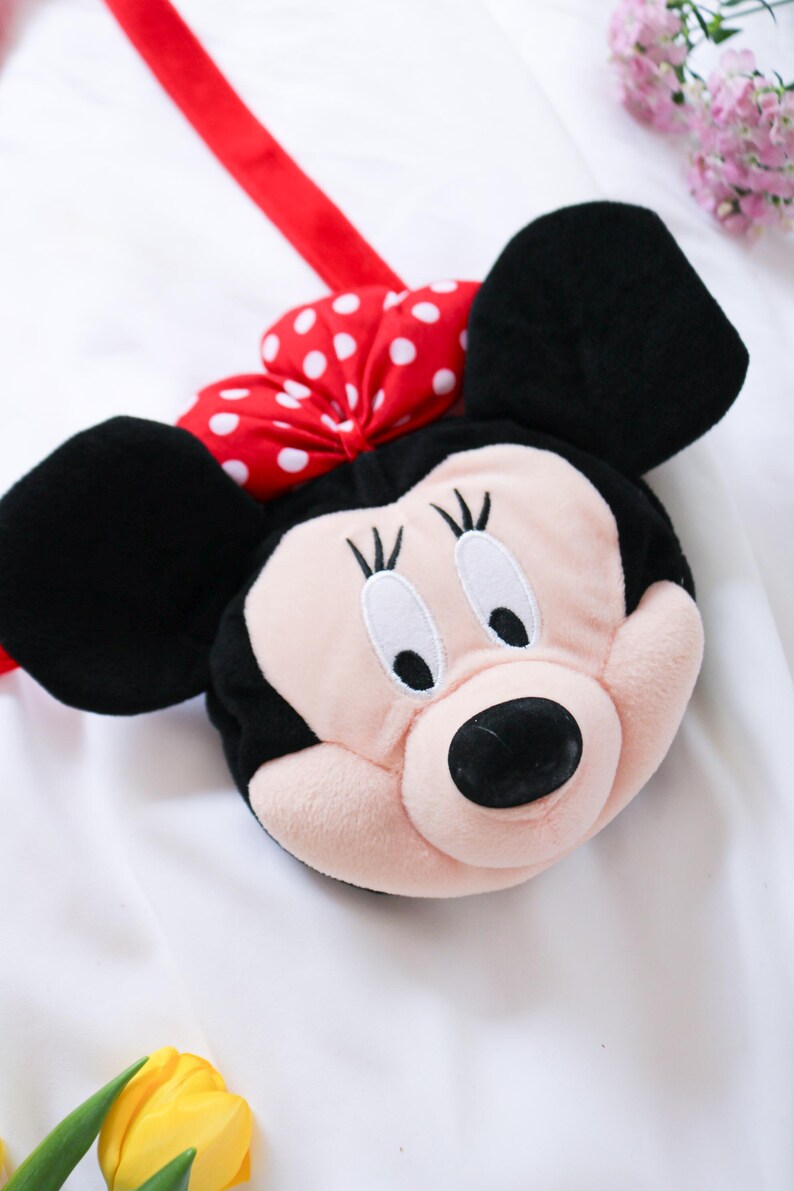 May include: A plush Minnie Mouse-shaped bag with a red strap. The bag features a pink face, black ears, and a red bow with white polka dots. The bag is designed to resemble the iconic Disney character.