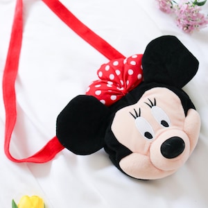 May include: A plush Minnie Mouse-shaped shoulder bag with a red strap and a red bow with white polka dots. The bag features black ears, a pink face, and a black nose. The bag is designed for children.