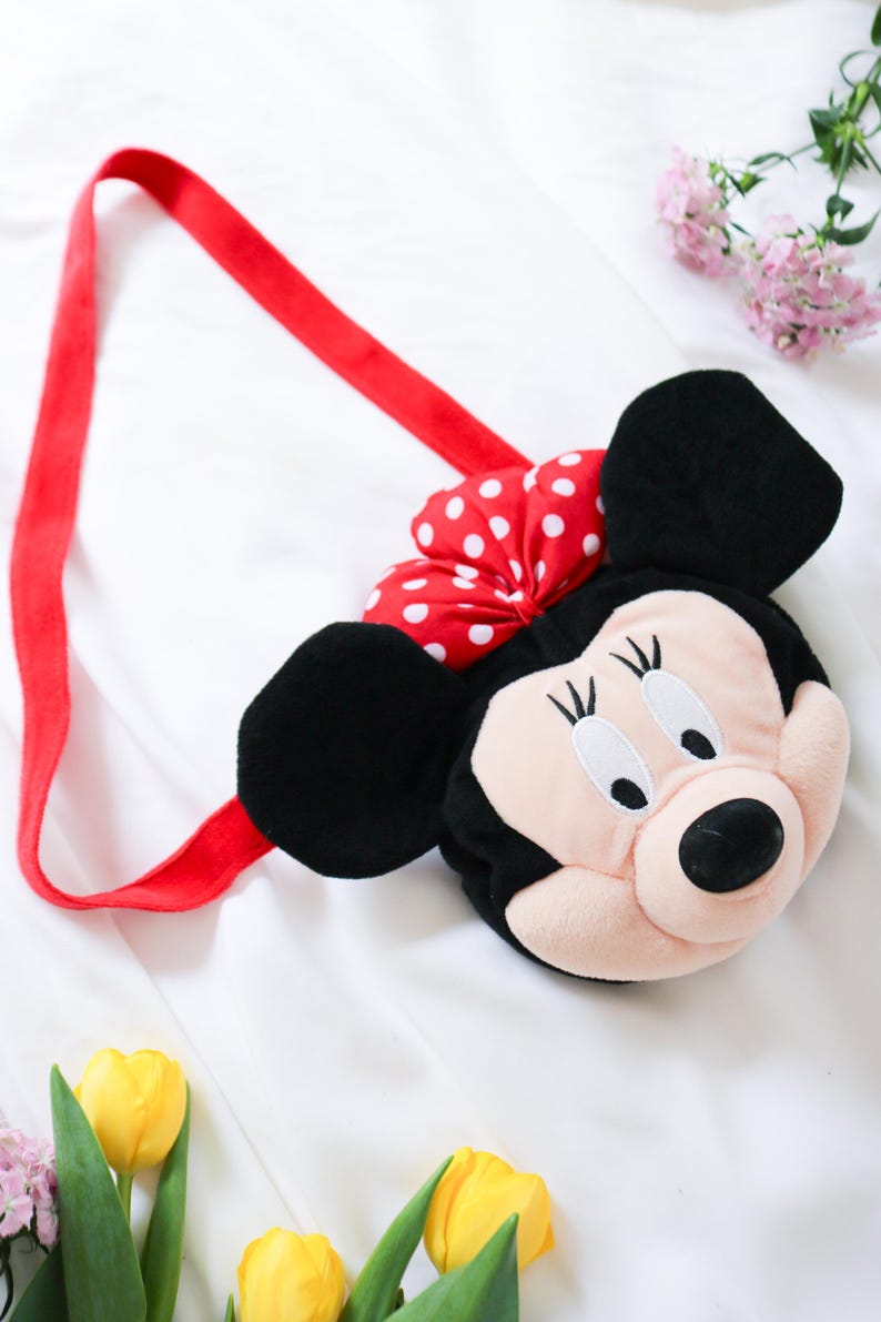 May include: A plush Minnie Mouse-shaped shoulder bag with a red strap and a red bow with white polka dots. The bag features black ears, a pink face, and a black nose. The bag is designed for children.