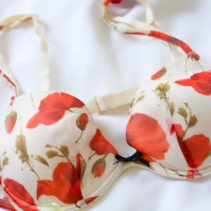 May include: A cream-colored bra with a vibrant red floral pattern. The bra features adjustable straps and a delicate design, with a black bow detail. The lingerie is set against a white background.