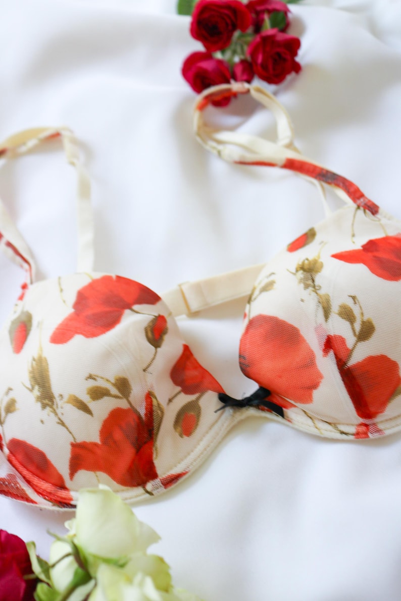 May include: A cream-colored bra with a vibrant red floral pattern. The bra features adjustable straps and a delicate design, with a black bow detail. The lingerie is set against a white background.