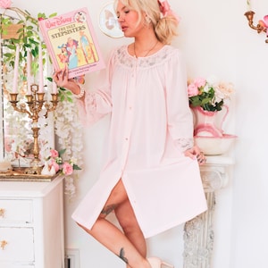 May include: A light pink vintage button-down robe with lace trim. The robe is knee-length and has long sleeves. The image also features a book titled "Walt Disney The Ugly Stepsisters".