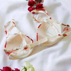 May include: A cream-colored bra with a floral pattern in red and green, featuring adjustable straps and a hook-and-eye closure. The bra is set against a white background with red roses.