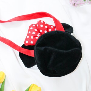 May include: Black plush handbag with a red strap and a red bow with white polka dots. The bag has two black ears and is designed to resemble a cartoon character. The bag is on a white background.