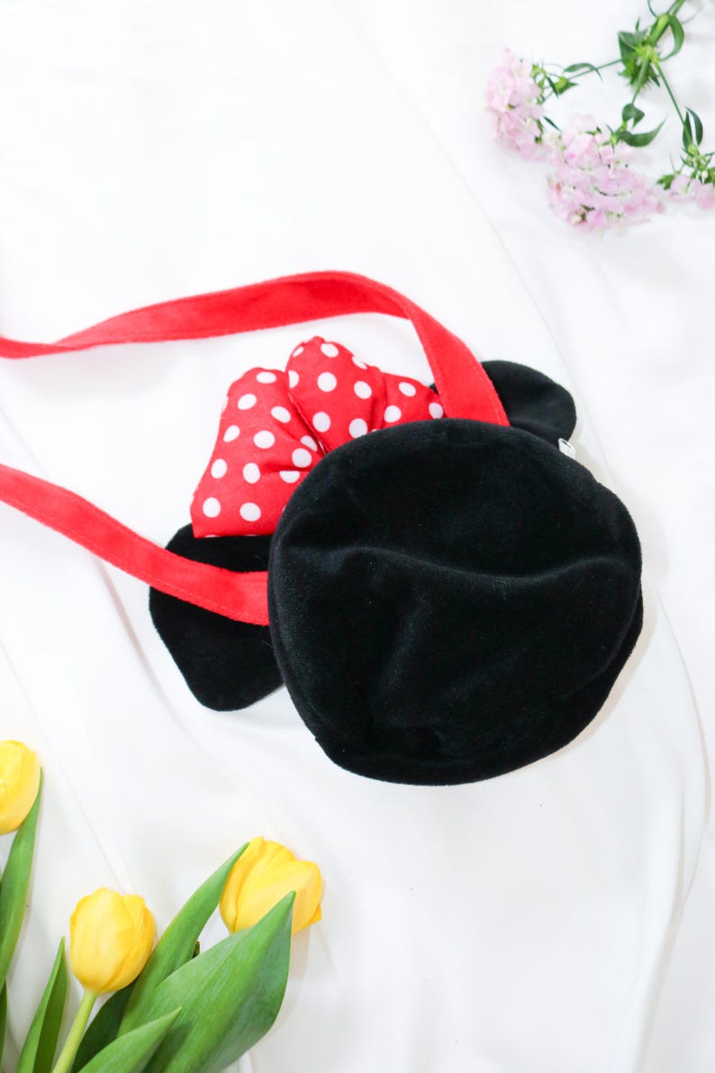 May include: Black plush handbag with a red strap and a red bow with white polka dots. The bag has two black ears and is designed to resemble a cartoon character. The bag is on a white background.