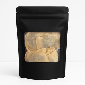 Puede incluir: Una bolsa negra con una ventana transparente que muestra un paquete envuelto en papel marrón claro. La bolsa tiene un cierre de cremallera. El embalaje está diseñado para contener alimentos y está sobre un fondo blanco.