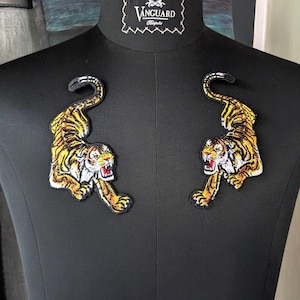 May include: Two iron-on patches featuring a yellow and black tiger design. The tigers are facing forward with their mouths open, showing their teeth. The patches are on a black background.