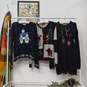 May include: Three sweaters hang on a white rack. The navy blue sweater features snowmen and snowflakes. The gray cardigan has bear and tree designs. The black sweater has colorful sequin designs. A framed botanical print hangs in the background.