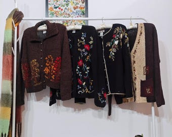 May include: A white metal clothing rack displays four vintage sweaters. The sweaters are various colors, including brown, black, and beige, and feature floral embroidery and patterns. A colorful scarf hangs on the rack's side.