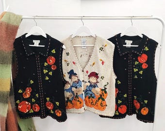 May include: Three sleeveless knit vests hanging on white hangers. Two black vests feature embroidered orange pumpkins and green vines. The center beige vest displays two scarecrow figures wearing hats, surrounded by pumpkins and autumn leaves.