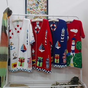 Op de afbeelding: Vier nautische cardigans in wit, rood, blauw en rood. Elke cardigan heeft een V-hals en korte mouwen. De ontwerpen omvatten zeilboten, vuurtorens, ankers en reddingsboeien. De cardigans hangen aan een wit rek.