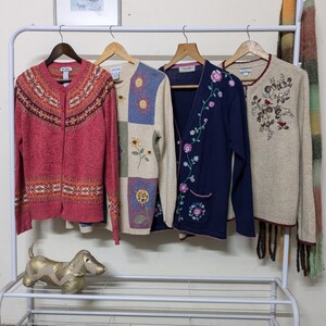 May include: Four different cardigans hanging on a white rack. The cardigan on the left is red with a white and orange pattern. The cardigan in the middle is cream with a patchwork design of flowers and squares. The cardigan on the right is beige with a floral pattern and a brown fringe. The cardigan in the centre is navy blue with a floral pattern.