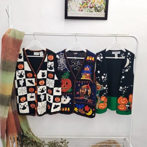 May include: Three sleeveless knit vests hanging on white hangers. The left vest features a black and white checkerboard pattern with orange pumpkins, ghosts, and bats. The middle vest is navy blue with a large pumpkin, spiderweb, and house. The right vest is navy blue with a green hem, stars, a moon, and pumpkins.