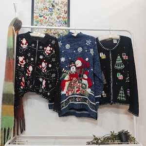 May include: Three festive Christmas sweaters are displayed on a white rail. A black zip-up sweater is decorated with Santa Claus motifs. A blue sweater features snowmen and snowflakes. A black cardigan has Christmas tree and gift box designs.