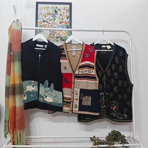 Könnte beinhalten: Ein Kleiderständer zeigt einen schwarzen Reißverschluss-Pullover mit Schafstickerei, eine Patchwork-Weste mit roten und blauen Einsätzen und eine schwarze Weste mit Blumenmuster. Ein farbenfroher Schal hängt links.