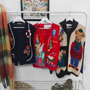 May include: Three vintage knit vests with bear and party-themed designs. The vests are navy, red, and beige, with embroidered details including bears, balloons, and presents. They hang on a white rack.