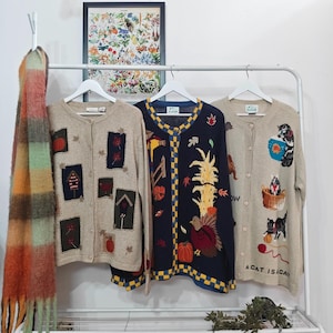 May include: Three knitted cardigans with autumn-themed designs hang on a white rail. One is beige with pumpkin and leaf patches, another is navy with a turkey and cornucopia, and the third is beige with cat graphics.