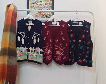 Könnte beinhalten: Drei Vintage-Westen hängen an einem weißen Gestell. Die Westen sind marineblau, weinrot und marineblau mit floralen und gartenartigen Stickereien. Ein farbenfroher Schal hängt links. Ein gerahmter botanischer Druck hängt darüber.