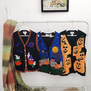 May include: Three sleeveless button-front vests hanging on white hangers. The left vest is black with ghosts and pumpkins. The middle vest is blue with a moon, tree, and pumpkins. The right vest is black with a pattern of orange jack-o'-lanterns.