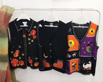 May include: Three sleeveless knit vests hanging on a white rack. The left vest is black with embroidered orange pumpkins. The middle vest is black with a pumpkin and moon design. The right vest features colorful Halloween patches with a witch and ghost.