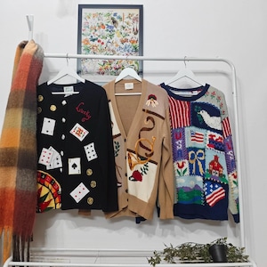 May include: Three vintage sweaters hang on a white rail. A black cardigan with playing card and gold coin designs, a tan cardigan with horn and floral embroidery, and a patchwork sweater with patriotic motifs.