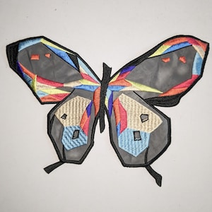 May include: A colourful geometric butterfly patch with a grey background and black outline. The butterfly has red, blue, yellow, and orange accents.