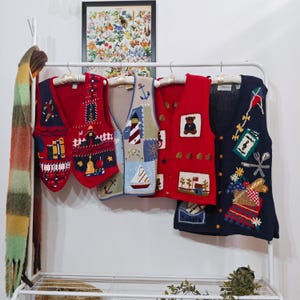 May include: A collection of colorful, vintage-style knit vests with various nautical and Americana-themed designs, including anchors, sailboats, and a lighthouse. The vests are displayed on a white rack with a scarf.