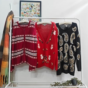 May include: Three sweaters hang on a white rail. A burgundy zip-up sweater with a white geometric pattern, a red cardigan with bear and floral embellishments, and a black sweater with gold sequin paisley designs.