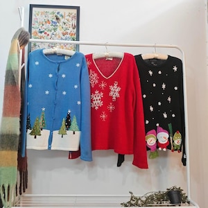 May include: Three Christmas sweaters hang on a white rack. A blue cardigan with snowflake and tree designs, a red V-neck sweater with snowflakes, and a black sweater with stars and snowman figures.