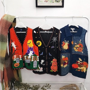 May include: Three sleeveless vests hanging on white hangers. The left vest is orange and green with ghosts, a haunted house, and tombstones. The middle vest is black with a spiderweb, bats, and pumpkins. The right vest is blue with autumn harvest motifs.