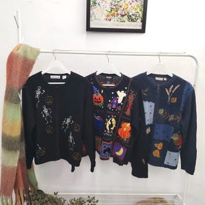 Puede incluir: Tres cárdigans de manga larga con botones en perchas blancas. El de la izquierda es negro con esqueletos blancos y calabazas naranjas. El central es negro con motivos coloridos de Halloween. El de la derecha es azul con patrones florales.