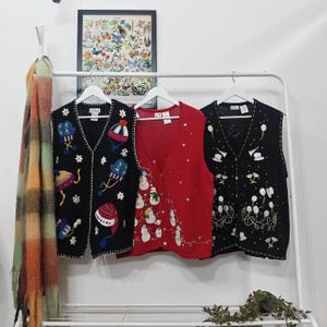 May include: Three festive vests hang on a white rail. The vests are black and red, with embroidered holiday designs. One black vest has colourful winter-themed designs, the red vest has snowmen, and the other black vest has champagne glasses and balloons.