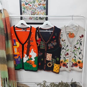Può includere: Tre gilet senza maniche con bottoni appesi a grucce bianche. Il gilet a sinistra è arancione con una casa stregata, fantasmi e zucche. Il gilet centrale è nero con motivi patchwork e girasoli. Il gilet a destra è beige con uccelli e fiori.