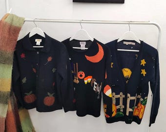 May include: Three dark navy blue sweaters hanging on white hangers. The left sweater features embroidered pumpkins and leaves. The center sweater displays a crescent moon, pumpkin, and candy corn. The right sweater shows a moon, stars, and pumpkins.
