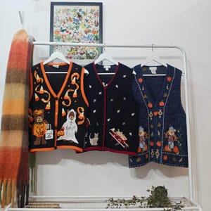 May include: Three decorative vests hang on a white rail. The vests are black, navy, and orange, with Halloween and autumn-themed designs. One vest has a bear holding a pumpkin, another has a ghost, and the third has scarecrows and pumpkins.