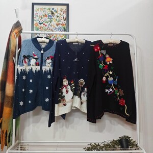 May include: Three Christmas-themed zip-up cardigans hanging on a white rail. The cardigans are blue, navy, and black, each with festive designs including penguins, snowmen, and gift boxes. The cardigans are made of a knitted material.