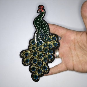May include: A green and gold embroidered peacock patch with a red crown. The peacock's tail feathers are spread out in a fan shape.