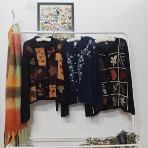 Op de afbeelding: Drie vintage vesten hangen aan een witte kledingrek. De vesten zijn zwart, marineblauw en zwart met blad- en bloemmotieven. Een kleurrijke sjaal hangt aan de linkerkant en een ingelijste botanische prent staat op de achtergrond.
