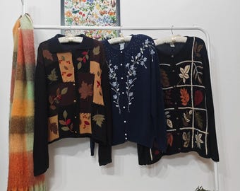 Könnte beinhalten: Drei Vintage-Strickjacken hängen an einem weißen Kleiderständer. Die Strickjacken sind schwarz, marineblau und schwarz mit Blatt- und Blumendesigns. Ein farbenfroher Schal hängt links, und ein gerahmter botanischer Druck befindet sich im Hintergrund.
