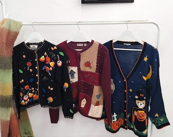 May include: Three long-sleeved button-down cardigans hanging on white hangers. The left is black with embroidered pumpkins and candy corn. The center is maroon with a patchwork design of birdhouses and pumpkins. The right is navy with a bear graphic.