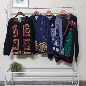 May include: Four Christmas sweaters hanging on a white rack. The sweaters are black, blue, and green with various holiday designs, including reindeer, snowmen, and Christmas trees.