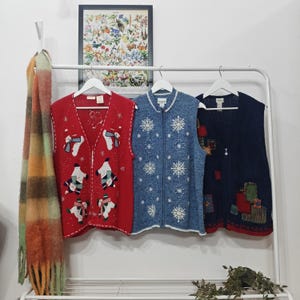 May include: Three vintage festive waistcoats hang on a white rail. A red one has snowman and snowflake designs. A blue one features snowflake patterns, and a navy one has patchwork details. A colourful scarf hangs on the left.