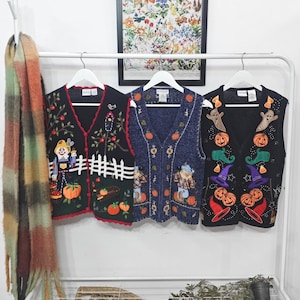 Peut inclure: Trois gilets en tricot sans manches suspendus sur des cintres blancs. Le gilet de gauche est noir avec un motif d'épouvantail et de citrouilles. Le gilet central est bleu avec des tournesols et des citrouilles. Celui de droite est noir avec des fantômes, des citrouilles et des chapeaux de sorcière.