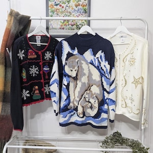 May include: Three sweaters hang on a rack. A black cardigan with Christmas-themed designs, a navy blue sweater with polar bears, and a cream-colored zip-up sweater with gold embroidery. The sweaters are displayed against a white wall.